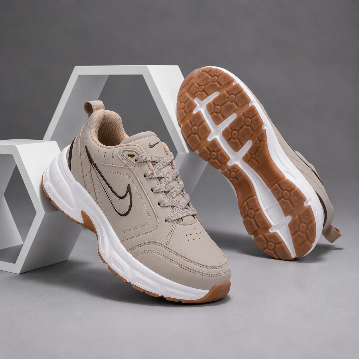A pair of chunky low-top sneakers in a light taupe-beige colorway, with one shoe standing upright and the other lying on its side to show the outsole. The shoes feature a dark brown swoosh logo, a thick white midsole, and a brown gum outsole, set against a gray background with white geometric display blocks.